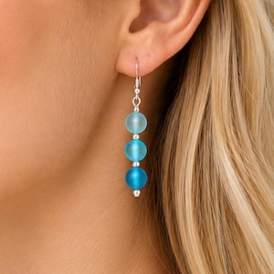 Può includere: Un orecchino pendente con tre perline sferiche disposte in un gradiente verticale di blu, che sfuma da un azzurro chiaro satinato in alto a un blu intenso in basso, separate da piccole perline distanziatrici in metallo color argento.
