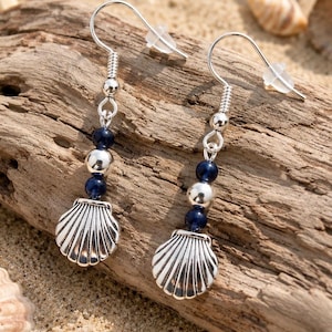 May include: Silver seashell earrings with blue and silver beads. The earrings have a fish hook style and are displayed on a piece of driftwood. The earrings are a beach-themed accessory.