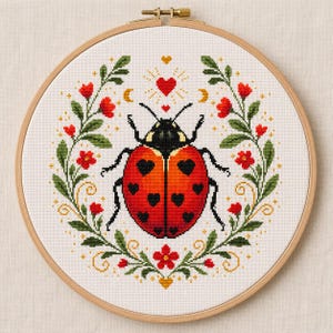Può includere: Un telaio da ricamo circolare in legno contenente un motivo a punto croce di una coccinella rossa con macchie nere a forma di cuore. La coccinella è centrata all'interno di una ghirlanda simmetrica di foglie verdi, fiori rossi e piccole stelle.