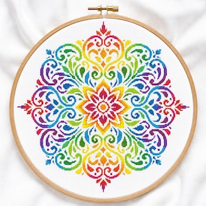 May include: A circular wooden embroidery hoop containing a cross-stitch pattern on white fabric. The design features a symmetrical, multi-colored mandala-style floral motif with vibrant red, orange, yellow, green, blue, and purple threads.