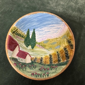 May include: A round ceramic plate with a hand-painted landscape scene. The design features a blue sky, green hills, trees, and buildings with red roofs. The plate has a brown rim and a rustic, artistic style.