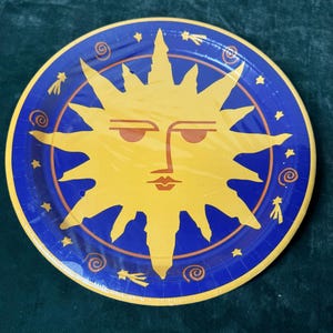 May include: A round paper plate with a yellow sun design in the center. The sun has a face with orange eyebrows and a mouth. The plate has a blue border with star and swirl designs.