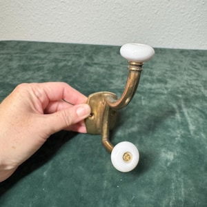 May include: Vintage brass wall hook with two white ceramic knobs. The hook has a curved design and a rectangular brass base. The knobs have a rounded shape and a gold-colored center. The hook is held by a hand.