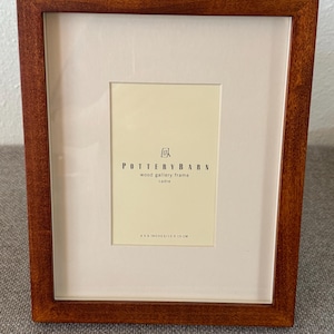 May include: A rectangular wooden picture frame with a warm brown finish. The frame has a cream-colored mat and a central opening. The text "POTTERY BARN wood gallery frame cadre" is printed on the mat. The frame is 4 x 6 inches / 10 x 15 cm.
