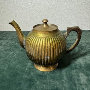 May include: A vintage brass teapot with a ribbed body, a curved spout, and a handle. The lid is topped with a small knob. The teapot sits on a round base. The brass has a warm, aged patina.
