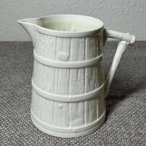 May include: A white ceramic pitcher with a barrel design. The pitcher has a spout, a handle, and horizontal bands around the body. The exterior is textured to resemble wooden planks. A classic kitchenware item.