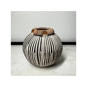 May include: A spherical vase featuring a black body with vertical white textured stripes and a rim wrapped in brown coiled material. The vessel has a rounded shape with a narrow opening at the top, showcasing a contrast between the dark base and light vertical lines.