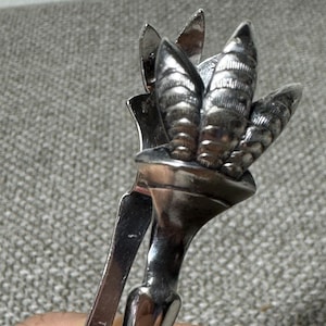 May include: A silver-toned metal sugar tong featuring a decorative top with three textured, leaf-like prongs. The handle is engraved with the text TRIPLE PLATE. The metal surface has a polished finish with visible reflections and intricate detailing.