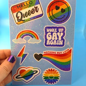 Può includere: Un foglio di adesivi con disegni olografici tra cui un cartellino Hello My Name Is Queer, una faccina sorridente arcobaleno con occhi a cuore, un arcobaleno, il testo Woke Up Gay Again, un fulmine, un cuore con scia arcobaleno e un fiore.