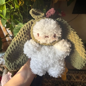 Puede incluir: Un peluche que representa una criatura redonda, blanca y esponjosa con un rostro bordado sencillo, que lleva una capucha de ganchillo de color verde oliva oscuro con orejas largas y caídas y un pequeño detalle rojo oscuro en la parte superior.