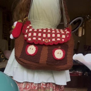May include: A brown crocheted messenger bag featuring a red and white chequered flap with a central wooden button. The bag includes two front pockets adorned with circular apple motifs and a small crocheted apple charm attached to the strap.