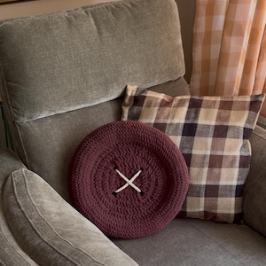 May include: A round, dark burgundy crochet pillow shaped like a large button. The pillow features a thick, textured knit surface with a central white cord stitched in an X pattern through four distinct holes, mimicking a functional garment button.