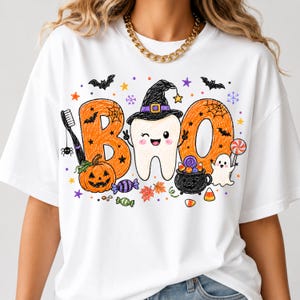 K&ouml;nnte beinhalten: Ein wei&szlig;es T-Shirt mit einem Grafikdruck des Wortes BOO, bei dem der Buchstabe B orange ist, der Buchstabe O durch einen zwinkernden Zahn mit Hexenhut ersetzt wird und das letzte O orange ist. Enth&auml;lt eine Zahnb&uuml;rste, K&uuml;rbis, Geist und S&uuml;&szlig;igkeiten.