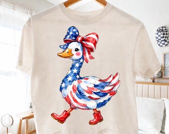 May include: Cream-colored t-shirt featuring a patriotic goose design. The goose is adorned with a red, white, and blue bow, and its body is painted with the American flag. It wears red boots and is walking.