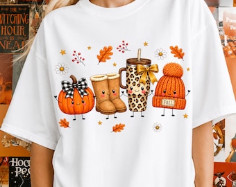 May include: A white t-shirt featuring a graphic of four anthropomorphic fall items: an orange pumpkin with a black and white checkered bow, tan winter boots, a leopard print tumbler with a gold bow, and an orange knit beanie labeled FALL.