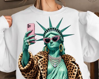 May include: White sweatshirt featuring a graphic of the Statue of Liberty wearing pink sunglasses, a leopard print coat, and jewelry, taking a selfie with a pink phone. The Statue of Liberty is depicted in a teal color. The design is a digital download.
