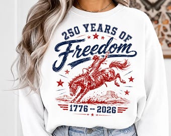 May include: White sweatshirt with a graphic design featuring a cowboy riding a bucking bronco. The design includes the text "250 Years of Freedom" and the years "1776-2026" in red and blue. The sweatshirt has a crew neck.
