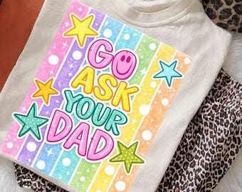 May include: A cream-colored t-shirt with a colorful graphic that reads "GO ASK YOUR DAD." The graphic features a smiley face and star shapes in various colors. Leopard print pants are visible in the background.