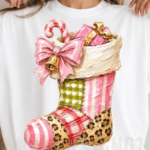May include: A white t-shirt featuring a textured, painted-style graphic of a Christmas stocking. The stocking has a cream cuff, a pink bow with a gold bell, a candy cane, and a gift. The body features green gingham, pink stripes, and leopard print.