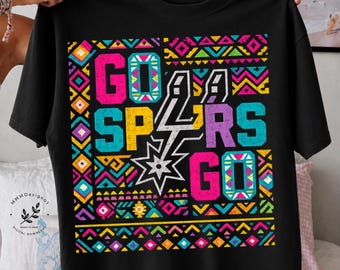 Puede incluir: Camiseta negra con un diseño geométrico colorido y las palabras "GO SPURS GO" en rosa brillante, amarillo, turquesa y morado. El logotipo de los Spurs está en el centro. El diseño está enmarcado por un borde vibrante.