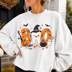 K&ouml;nnte beinhalten: Ein wei&szlig;es Langarm-Sweatshirt mit einem Grafikdruck, der das Wort BOO bildet. Der Buchstabe B ist orange mit einer Zahnb&uuml;rste und einem K&uuml;rbis, der Buchstabe O ist ein zwinkernder Zahn mit Hexenhut, und das zweite O ist orange mit Geist und Kessel.