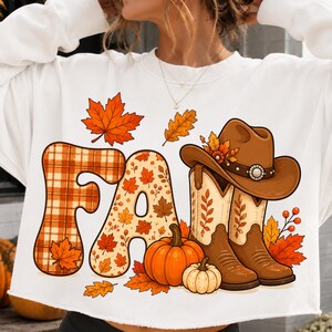 Könnte beinhalten: Ein weißes Langarm-Sweatshirt mit einem Grafikdruck des Wortes FALL. Der Buchstabe F hat ein orangefarbenes Karomuster und der Buchstabe A ist mit Herbstblättern gefüllt. Ein brauner Cowboyhut, zwei braune Cowboystiefel und zwei Kürbisse.