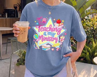 May include: A blue short-sleeved t-shirt with the text "Teaching is my Ministry" inside a star graphic. The graphic includes a pencil, apple, books, and other school-related icons. The person is holding a clear plastic cup with iced coffee.