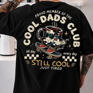 Pode incluir: Camiseta preta com um desenho de uma xícara de café usando óculos de sol em um skate. O texto diz "PROUD MEMBER OF THE DADS CLUB" e "STILL COOL JUST TIRED". O design inclui estrelas e padrões quadriculados.