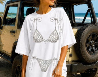 May include: White t-shirt featuring a rhinestone bikini top and bottom design. The design includes bows and dangling rhinestone accents. The shirt is worn outdoors near a beach and a vehicle with surfboards on top.