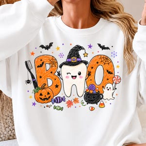 K&ouml;nnte beinhalten: Ein wei&szlig;es Sweatshirt mit Rundhalsausschnitt und einer bunten Grafik des Wortes BOO, in dessen Mitte ein Zahn mit Hexenhut abgebildet ist. Das Design zeigt einen K&uuml;rbis, eine Zahnb&uuml;rste, einen Kessel, ein Gespenst, S&uuml;&szlig;igkeiten, Flederm&auml;use und Sterne.