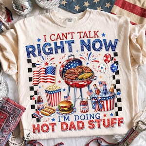 May include: Cream-colored t-shirt with a patriotic barbecue design. The shirt features the text "I CAN'T TALK RIGHT NOW I'M DOING HOT DAD STUFF" with images of a grill, food, and American flags. The shirt is decorated with red, white, and blue colors.