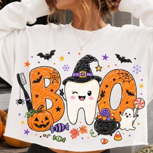 K&ouml;nnte beinhalten: Ein wei&szlig;es Langarm-Sweatshirt mit einem bunten Grafikdruck und dem Schriftzug BOO. Das Design zeigt einen zwinkernden Zahn mit Hexenhut, einen orangefarbenen K&uuml;rbis, einen schwarzen Kessel voller S&uuml;&szlig;igkeiten, ein Gespenst und Flederm&auml;use.