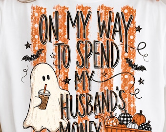 May include: A white t-shirt featuring a graphic with orange vertical stripes and the text ON MY WAY TO SPEND MY HUSBAND'S MONEY. The design includes a ghost holding an iced coffee, two flying bats, and a shopping cart filled with various pumpkins.