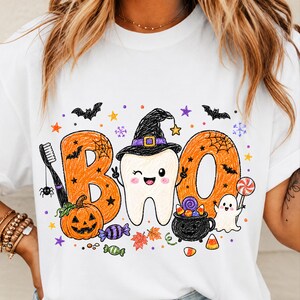 K&ouml;nnte beinhalten: Ein wei&szlig;es T-Shirt mit einem Grafikdruck des Wortes BOO in Orange, mit einem zwinkernden Zahn in der Mitte, der einen schwarzen Hexenhut tr&auml;gt. Das Design umfasst eine Zahnb&uuml;rste, einen K&uuml;rbis, einen schwarzen Kessel, ein Gespenst und Halloween-Symbole.