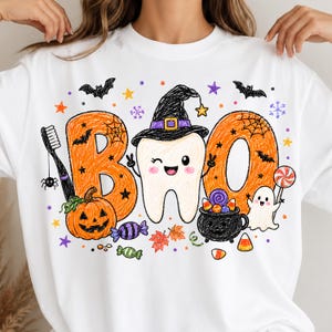 K&ouml;nnte beinhalten: Ein wei&szlig;es Sweatshirt mit Rundhalsausschnitt und einer bunten Grafik im Buntstift-Stil mit dem Text "BOO". Das Design zeigt einen zwinkernden Zahn mit Hexenhut, einen orangefarbenen K&uuml;rbis, einen schwarzen Kessel, ein Gespenst, eine Zahnb&uuml;rste und S&uuml;&szlig;igkeiten.