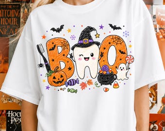 May include: A white t-shirt featuring a graphic print of the word BOO with a winking tooth wearing a witch hat in the center. The design includes a toothbrush, a jack-o'-lantern, a cauldron with candy, a ghost, bats, stars, and colorful autumn leaves.