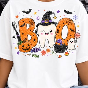 K&ouml;nnte beinhalten: Ein wei&szlig;es T-Shirt mit einer bunten Grafik und dem Schriftzug BOO, wobei der Buchstabe B orange ist, der Buchstabe O ein zwinkernder Zahn mit schwarzem Hexenhut ist und das zweite O orange ist. Enth&auml;lt eine Zahnb&uuml;rste, K&uuml;rbis, Geist und S&uuml;&szlig;igkeiten.