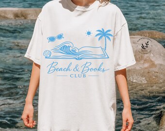 May include: White t-shirt with a light blue graphic. The graphic features sunglasses, a seashell, waves, an open book, a sun, and a palm tree. The text "Beach & Books Club" is below the graphic.
