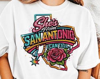 May include: White t-shirt featuring a colorful graphic design. The design includes the text "She's from San Antonio I can tell" in a stylized font, along with a rose and a star. The design is set within the outline of the state of Texas.