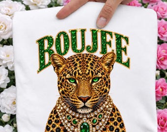 May include: White sweatshirt featuring a leopard design with green eyes and a jeweled necklace. The word "BOUJEE" is printed above the leopard in green glitter lettering. The sweatshirt is held up by a hand.