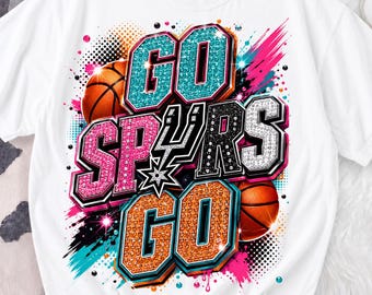 May include: White t-shirt with a vibrant graphic. The design displays "GO SPURS GO" in sparkling teal, pink, orange, and black lettering, with basketballs and a star. The shirt has a crew neck and short sleeves.