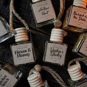 May include: An assortment of car air fresheners with wooden tops and labels. The labels feature text such as "Witch's Cottage", "Hexes & Honey", and "Burned at the Stake". Each bottle is suspended by a natural rope.