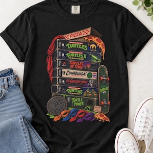 May include: A black t-shirt featuring a graphic print of a stack of VHS tapes labeled with Teenage Mutant Ninja Turtles titles, a pizza box, a skateboard, a manhole cover marked NYC SEWER, and four colored fabric masks.