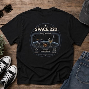 May include: A black t-shirt featuring a circular graphic on the back. The design displays the text SPACE 220, ELEVATED DRINKING, and Out of This World, with an illustration of three cocktail glasses against a view of Earth from space.