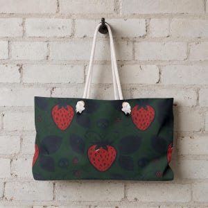 Puede incluir: Un bolso tote rectangular con fondo verde oscuro y un patrón repetitivo de fresas rojas, hojas oscuras y pequeñas calaveras. El bolso tiene dos asas de cuerda blanca sujetas con ojales de metal de tono plateado en el borde superior.