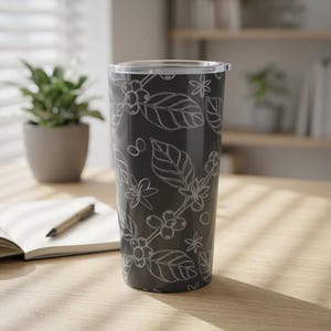 May include: A dark gray stainless steel tumbler featuring a repeating white line art pattern of coffee plant leaves, flowers, and coffee cherries. The cylindrical container has a clear plastic lid and a smooth, tapered body design.