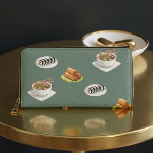 May include: A sage green rectangular zip-around wallet featuring a repeating pattern of Asian-inspired food illustrations, including bowls of ramen with chopsticks, plates of sushi rolls, and golden-brown fried spring rolls on lettuce leaves.