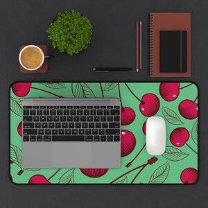 May include: A computer desk mat with a green background and a pattern of red cherries and leaves. A laptop, white mouse, and other office supplies are on the mat. A cup of coffee and a small plant are also visible.