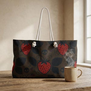 Puede incluir: Un bolso tote de color marrón oscuro con un patrón repetitivo de fresas rojas, hojas negras y pequeñas calaveras. El bolso tiene dos asas de cuerda blanca sujetas con ojales metálicos plateados. Una taza de cerámica beige se sitúa delante.