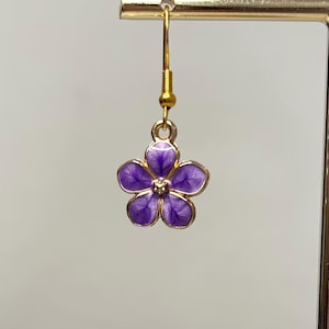 May include: A single gold-colored earring with a purple flower pendant. The flower has five petals with a gold outline and a small gold center. The earring is hanging from a gold hook.